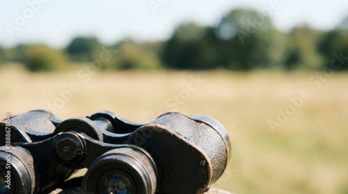 Weathered black binoculars with a textured grip stand in the foreground against a soft focus natural outdoor scene