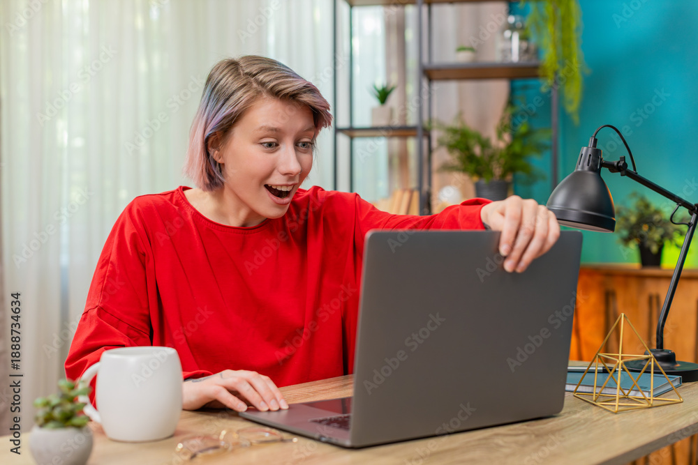 Fototapeta premium Young woman happily opening laptop sitting at home desk, celebrating excitedly after seeing lottery win message. Caucasian girl smiling joyfully, emotional cheerful reaction to big success at table.