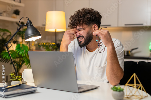 Arabian man at home feels headache and eye strain from screen holds head takes short break then. Egyptian guy sitting at table drinks water blinks rests breath calm returns to typing resumes work eve.