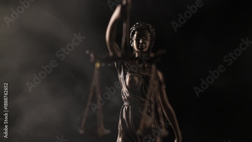 Wisps of white smoke drift slowly around the illuminated bronze figure of a blindfolded Lady Justice statue holding scales against a dark background.