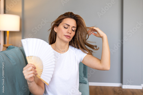 Tableau sur toile Young woman sitting on sofa using a white hand fan to cool down from hot flashes