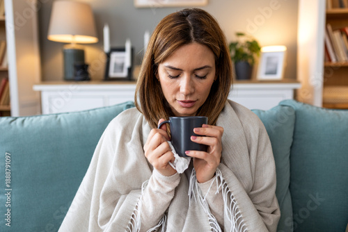 Fotografie Sick young woman wrapped in a blanket sitting on the sofa and drinking a cup of