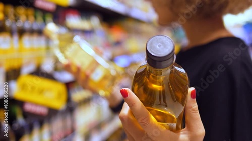 Close up of a young woman's hands with red manicure holding a glass bottle of olive oil, attentively comparing products and prices on the supermarket shelves before making her purchase