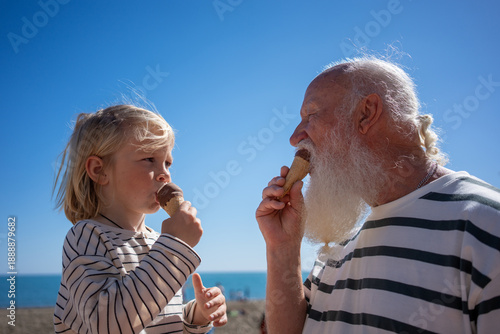 A heartwarming moment between a grandfather and his grandson as they share ice cream on a sunny beach day, smiling at each other with joy under a clear blue sky