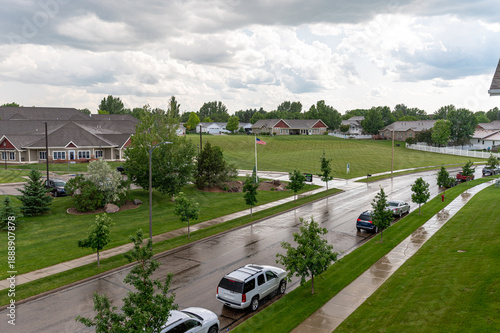 Quiet neighborhood in USA after rain, wet road with parked cars, green lawns, houses under cloudy sky.