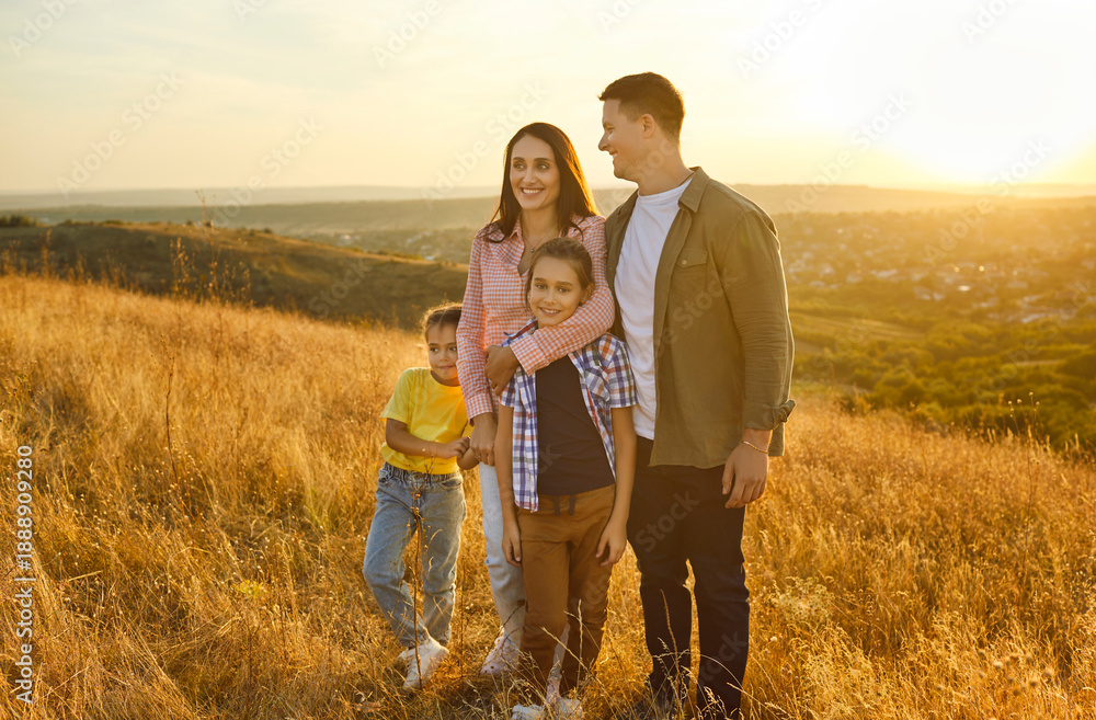 Fototapeta premium Happy family standing together at sunset, parents hug children. Father, mother, daughter and son enjoy golden light on meadow and hills in nature, calm moment of love and happiness of weekend hike
