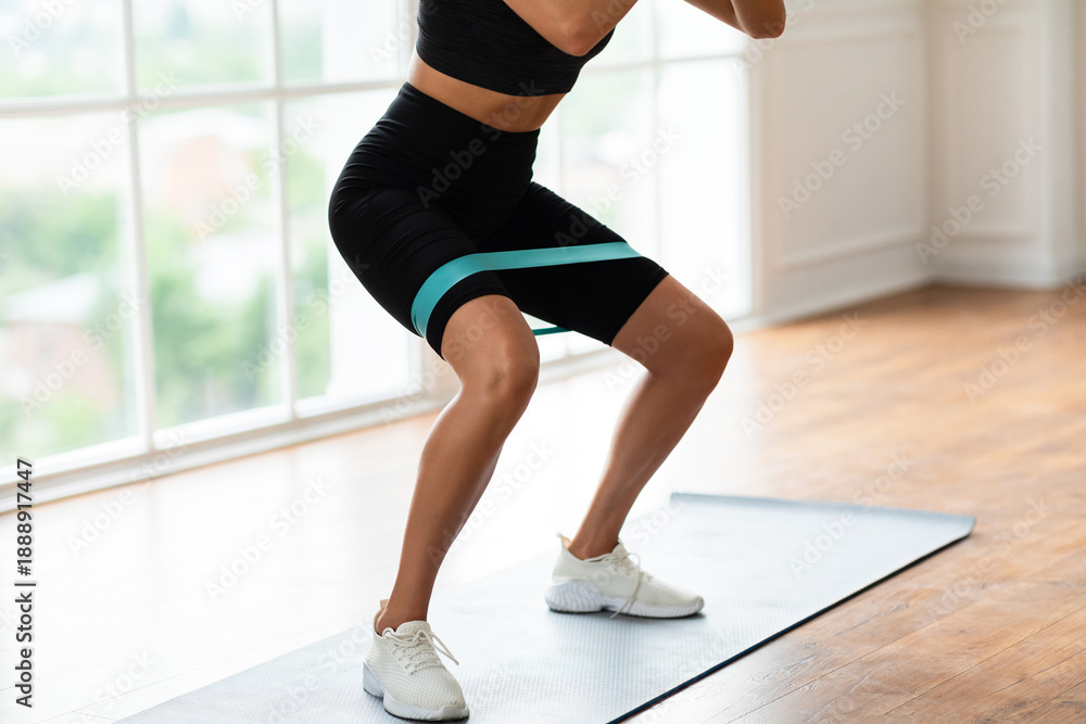 Fototapeta premium Closeup cropped view of athletic sportswoman squatting with elastic resistance band in fitness studio or at home in living room. Lady enjoying leg and buttocks work out with equipment
