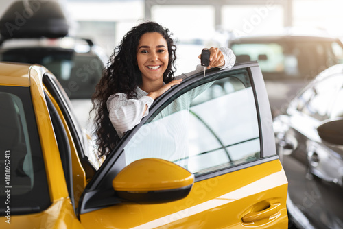 Cuadro en lienzo Cheerful brunette woman buying new cute car, raising hand up with automatic key and smiling, showroom interior, copy space