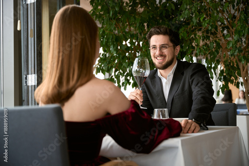 Happy young man in jacket with glass with wine looking at woman on the date in the restaurant