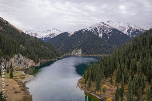 Wallpaper Mural Aerial view of a calm mountain lake surrounded by evergreen forest and alpine slopes, with snow-capped peaks in the background and reflections on the water. Torontodigital.ca
