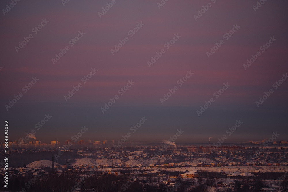 Naklejka premium Urban landscape features a vast expanse of snow-covered houses and buildings under a dramatic purple and blue sky during a cold winter sunset, highlighting the silhouette of the city horizon