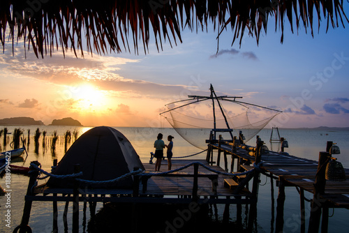 Overhead rear view of a woman sitting on a wooden jetty with two kayaks moored alongside at sunset, Ko Mat, Songkhla Lake, Phatthalung, Thailand
