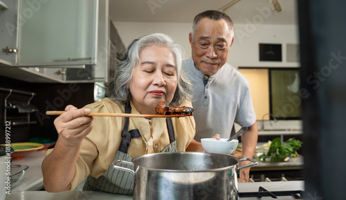 Two senior adults enjoy cooking together in a cozy kitchen. One offers a spoonful of food while the other leans in to smell, capturing warmth, sensory joy, and togetherness in a home lifestyle setting