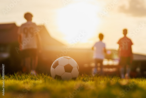 Soccer ball on green grass field with blurred silhouette of children playing football at sunset, golden hour light. Kids sport training concept.