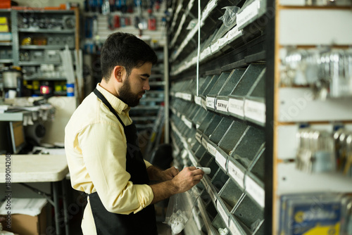 Canvas Print Young man employee wearing apron sorting small parts from storage bins into a ba