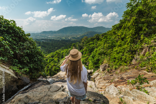 Fotografie Young woman wearing straw hat standing on rocky ledge overlooking lush tropical