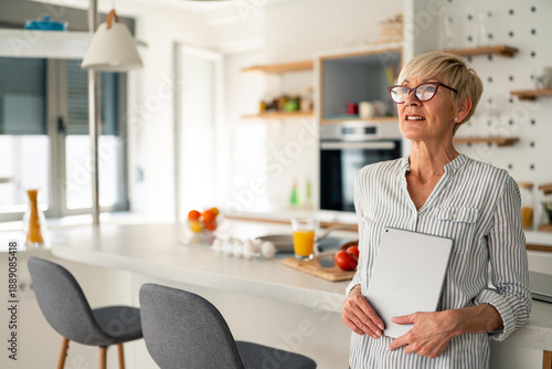 Billede på lærred Bright Morning Light Senior Woman Holding Laptop in Modern Kitchen Enjoying Work