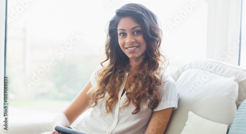 Billede på lærred Asian woman sitting on sofa at home by bright window holding dark tablet wearing