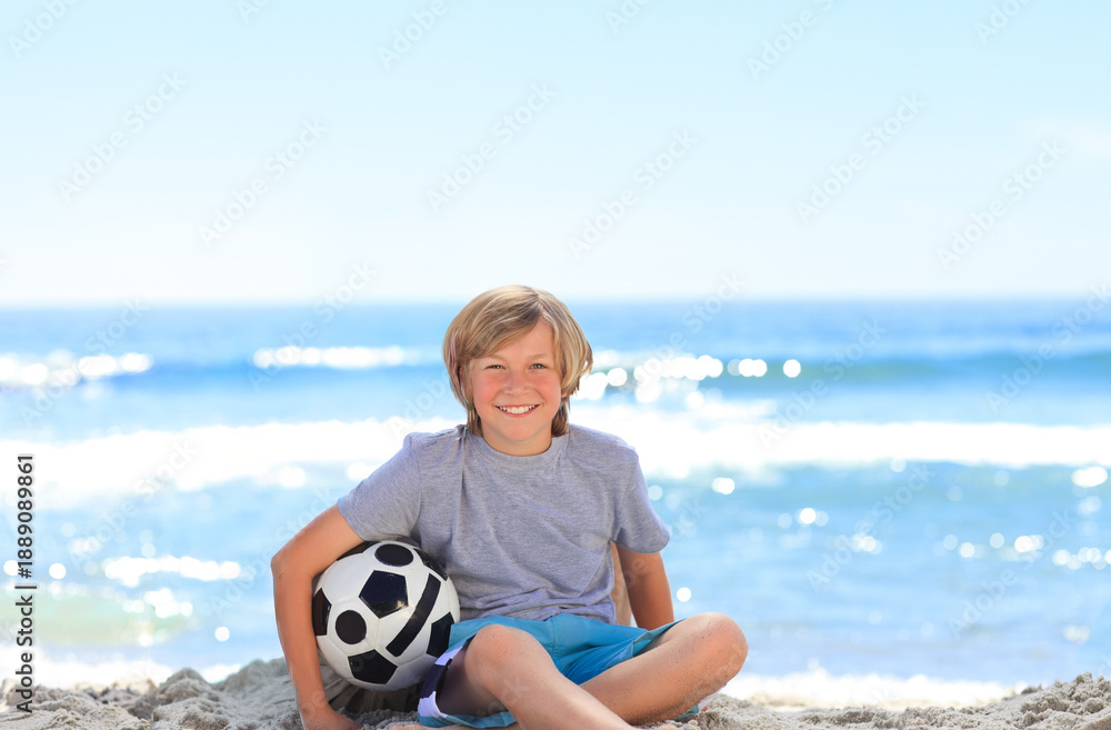 Fototapeta premium Child boy sitting cross-legged on sand holding black-and-white soccer ball at sunny beach