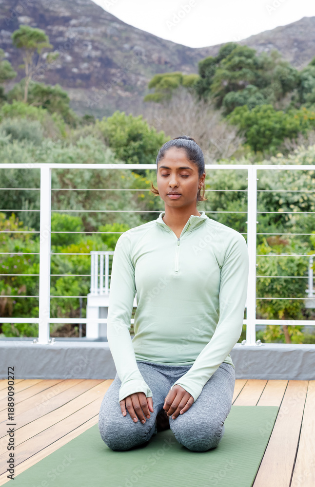 Fototapeta premium Indian woman kneeling on green yoga mat on wooden deck, eyes closed, in athletic wear