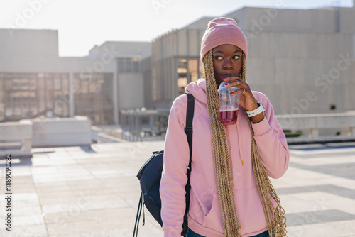 African American woman wearing pink beanie hoodie standing on rooftop sipping red drink, copy space