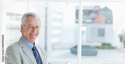 Senior man standing near glass windows in office in gray suit holding reading glasses, copy space