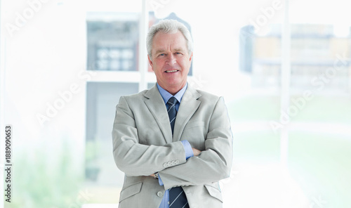 Senior man standing arms folded in bright lobby near windows wearing gray suit blue shirt tie