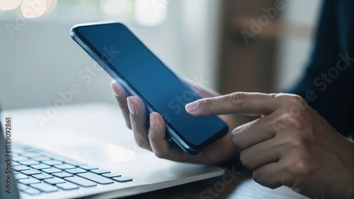 A person interacts with a smartphone while seated at a desk with a laptop, emphasizing technology and connectivity in a modern workspace.