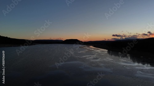 Aerial cinematic view of a calm lake at sunset, dark water surface reflecting the orange twilight sky, silhouettes of mountains and forests in the background, peaceful evening landscape