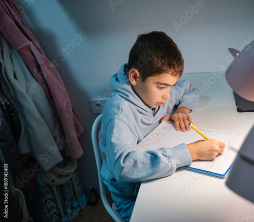 Young boy doing his homework alone at his desk in a cozy bedroom with warm lighting and focused expression during evening study time