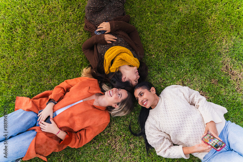 Three smiling women friends lying on green grass, heads together, enjoying time outdoors