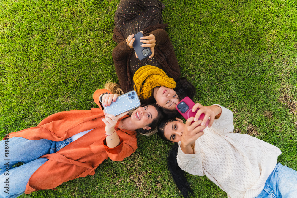 © pablo - Three young women friends lying on green grass taking selfies with smartphones