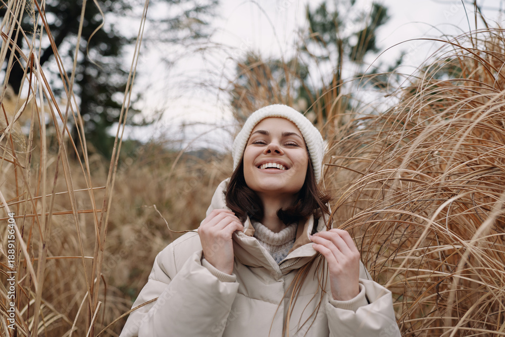 Fototapeta premium Woman in a warm beige coat and knit hat stands among tall grasses, smiling under a cloudy sky, capturing a cozy outdoor moment in late autumn or early winter soft breeze.