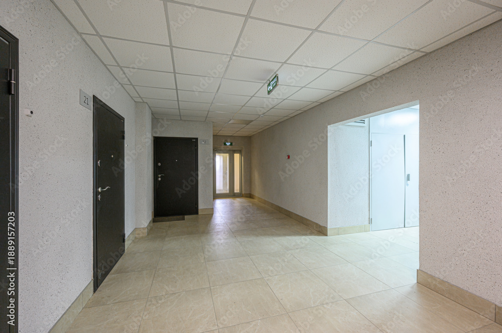 Fototapeta premium Long, light-toned corridor with tiled floor, textured walls, dark metal doors, white ceiling tiles, exit sign, clean, minimalist design, natural light at the end.