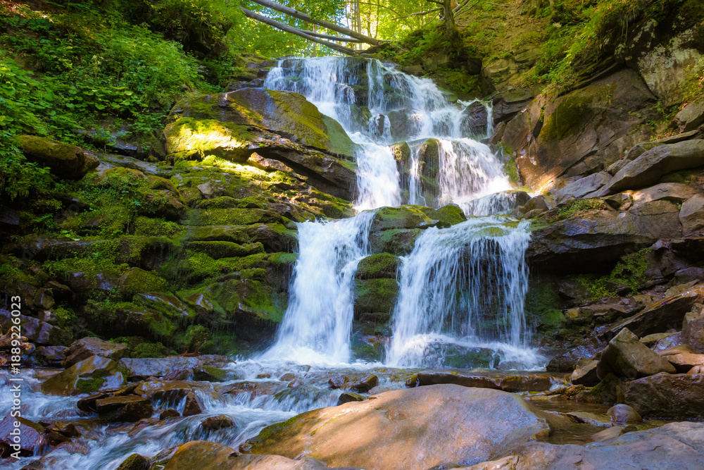 Obraz premium waterfall in the forest on a summer morning. rocks with moss in dappled light. refreshing nature scenery