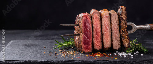 Classic five steak doneness degrees - Rare, Medium Rare, Medium, Medium well, Well done. Two-pronged fork piercing through stack of five steaks on dark stone surface with spices. Copy space