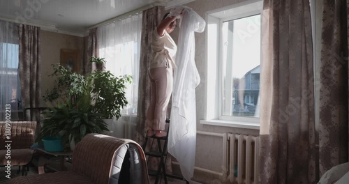 A housewife stands on a stepladder in the living room, removing white tulle curtains from the curtain rod for washing and cleaning. The woman is doing a general spring cleaning of the house.