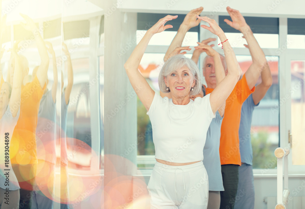 Fototapeta premium Beautiful mature women and sports men practice at ballet standing in fifth position at barre in bright choreography studio