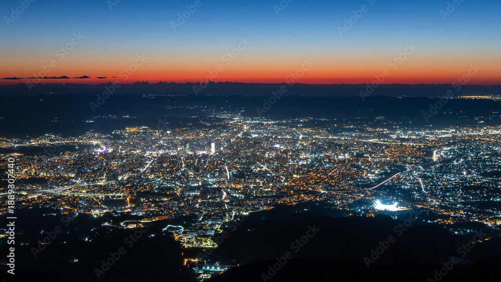 Naklejka premium Wide aerial view of a densely built capital illuminated after sunset with streets, neighborhoods and mountain silhouettes in sunset sky, Tirana, Albania