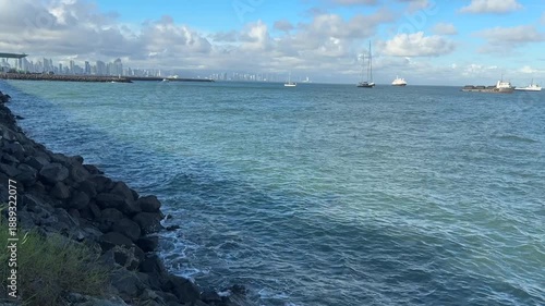 Several cargo ships and ferries anchored in Panama Bay, with shipping traffic on a sunny day against a cloudy backdrop