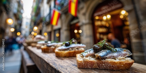 Spanish pintxos with anchovy, green olive, and red pepper on bread, served on narrow wooden bar counter, open to a stone alleyway with hanging flags and rustic village charm in background.