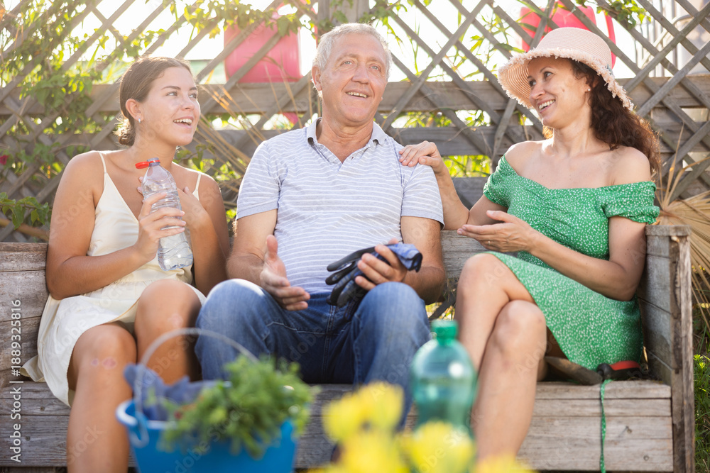 Fototapeta premium European woman in straw hat sitting on wooden bench, chatting sweetly with sister and elderly father. Rustic setting, young women and gray-haired man on background of wooden trellis discussing news
