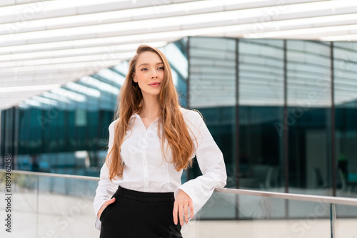 Free Download Professional young businesswoman in white shirt standing near railing with confident expression and modern glass building in background - GFXLoad