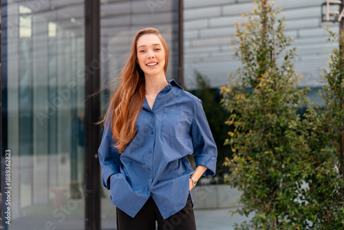 Smiling businesswoman in blue shirt standing near modern glass office building with confident and positive expression during daytime