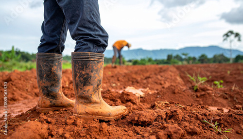 Muddy boots standing in a field with a person working in the background under a cloudy sky