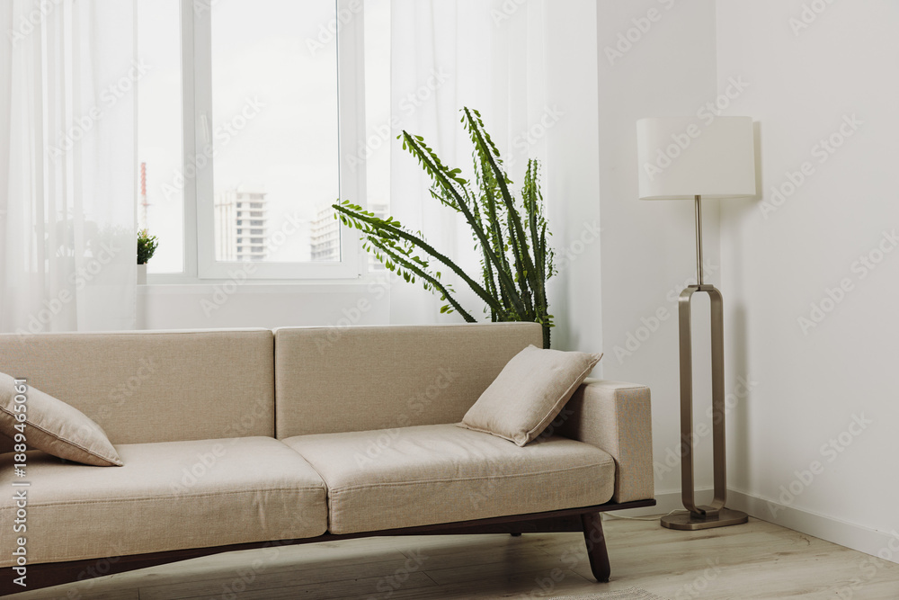 Fototapeta premium Minimalist living room with beige sofa and matching cushions under large bright windows. Tall green plant and modern floor lamp add natural and contemporary elements to the bright interior.