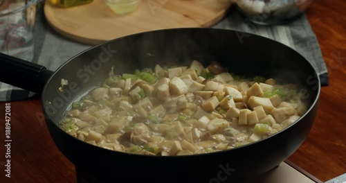 Housewife fries chopped mushrooms with onions in a frying pan, stirring them during home cooking. Close-up of homemade vegetarian food preparation in a cozy kitchen, natural light and authentic