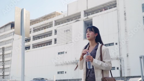 Young businesswoman walking outdoor street, shielding eyes from sun, holding water bottle, looking up at modern office buildings, enduring hot weather during city travel, professional career path.