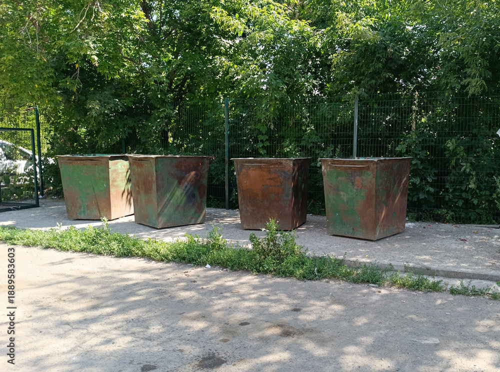 Naklejka premium Row of heavily rusted industrial trash bins sits in the bright sun, surrounded by lush green trees and a chain-link fence. Waste disposal