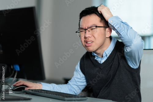 A confused young Asian businessman holds his hand to his head while working at his computer, ideal for workplace challenges, technical difficulties, or problem solving struggles.
